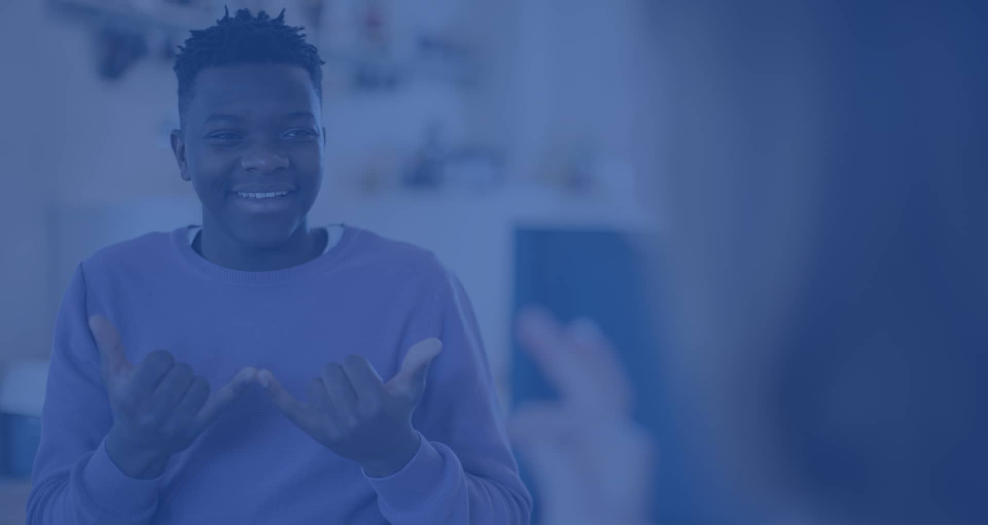 Black man in a purple sweater signing "now" in ASL to someone facing away from the camera.