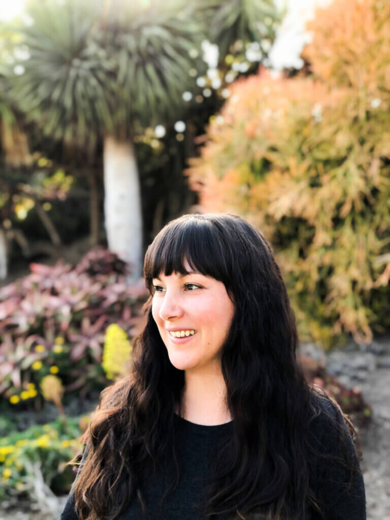 Brielle is light-skinned standing facing to their left, their long black hair with bangs goes past their shoulders laying onto their long black sweater. They're smiling away from the camera outside in a nature-like environment where tress of various shades of green can be seen with hints of purple too.
