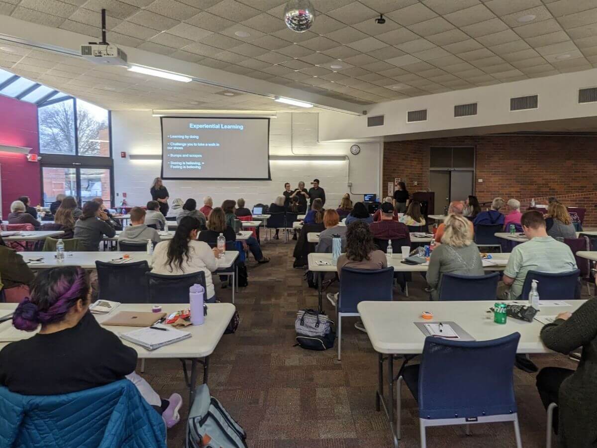 A shot from the back of a multipurpose room. The room is full of about 70 people sitting at tables. At the front of the room, the faces of Ashley Benton, Taylor Ofori, and the interpreting team are visible along with a PowerPoint slide entitled “Experimental Learning”.