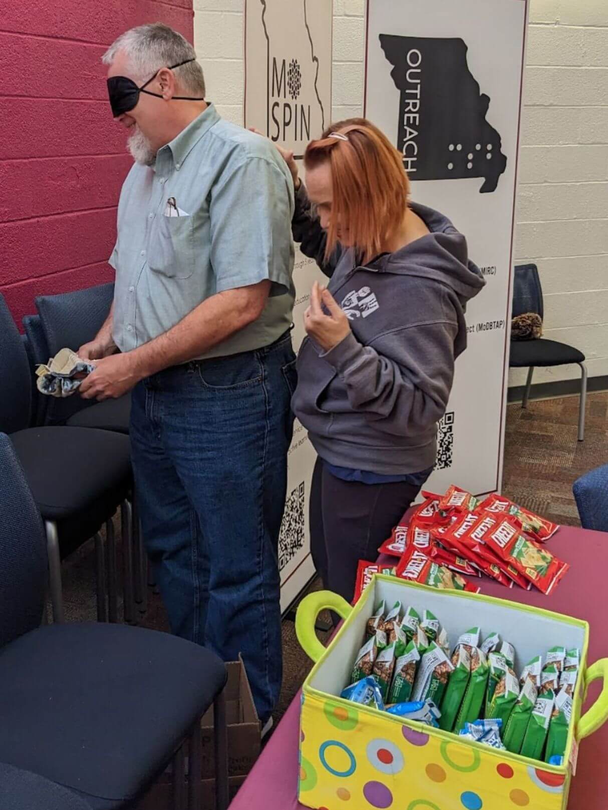 Two participants are involved in searching and finding for an object using haptic signals. In the foreground there are boxes of snacks, and in the background two tall signs explaining the MO DeafBlind Project are on display.