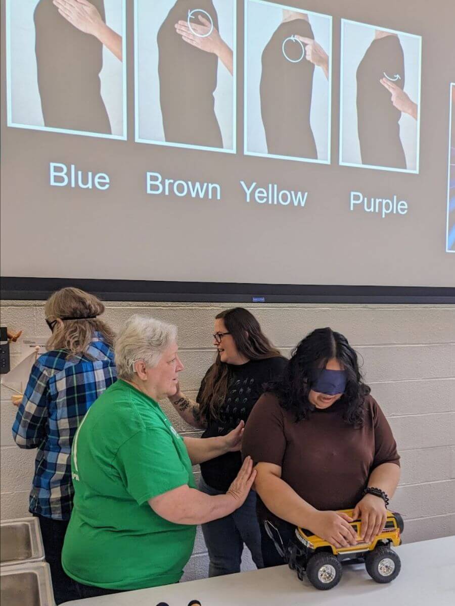 In the foreground, Mary Hale is using Haptic signals to describe a yellow truck to Vanessa Reyes, who is blindfolded. In the background, two more participants are involved in a similar activity and a PowerPoint slide displays the haptic signals for colors.