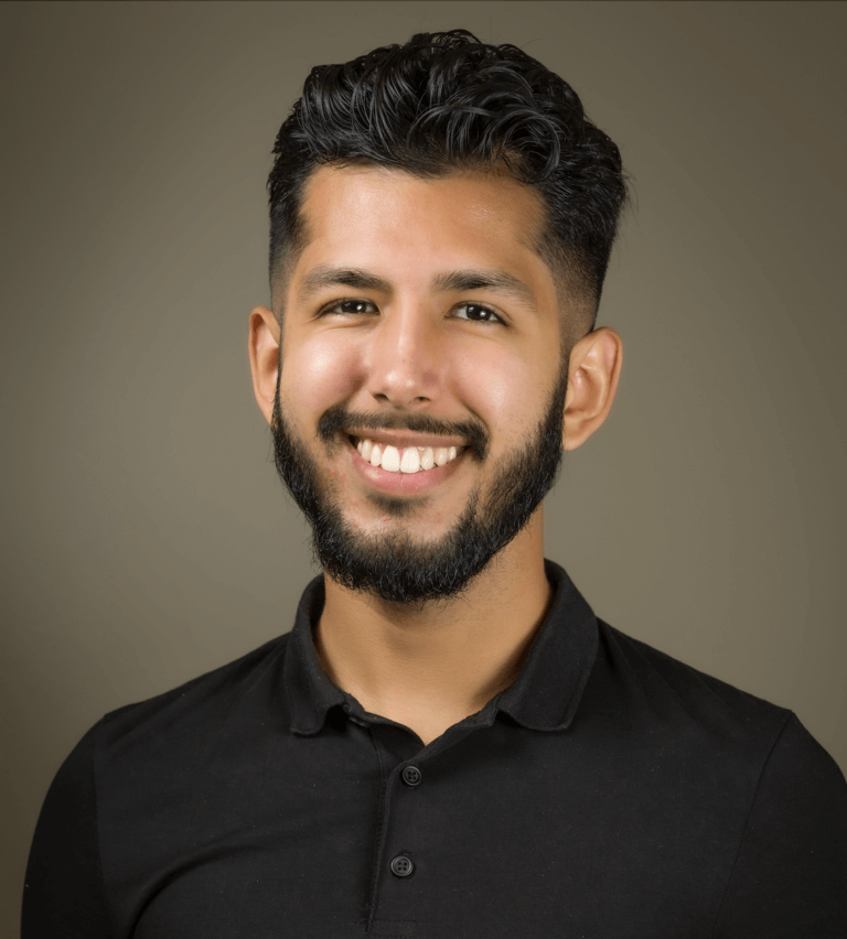 Headshot of Felix Villarreal who is tanned skinned with pushed back curly black hair and facial hair. Felix is smiling at the camera wearing a short-sleeved black polo shirt in front of a gray background.