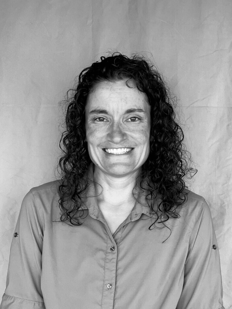 A black and white photo of Megan Cagno, smiling with long hair and wearing a buttoned up shirt against a plain background.
