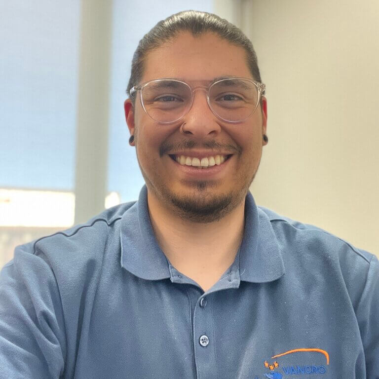 A photo of Mark Lucero-Muñiz, a man with dark facial hair and hair tied back wearing clear glasses and a blue Vancro polo shirt with a window and white wall in the background.