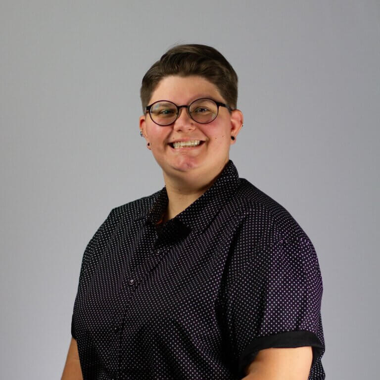 Photo of a white woman with short brown hair wearing glasses and a black button down shirt against a gray background.