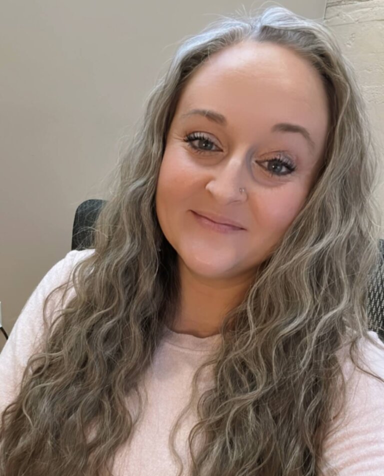 A photo of Jillian Watkins, a woman smiling with long hear and wearing a white long sleeved shirt against a white wall in the background.