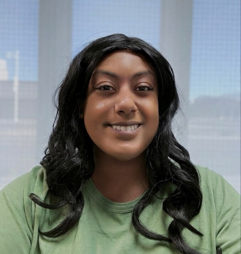 A photo of Lydia Tesfaye, a person with long black hair wearing a green shirt standing in front of a background that has a skyline and buildings blocked by blinds.