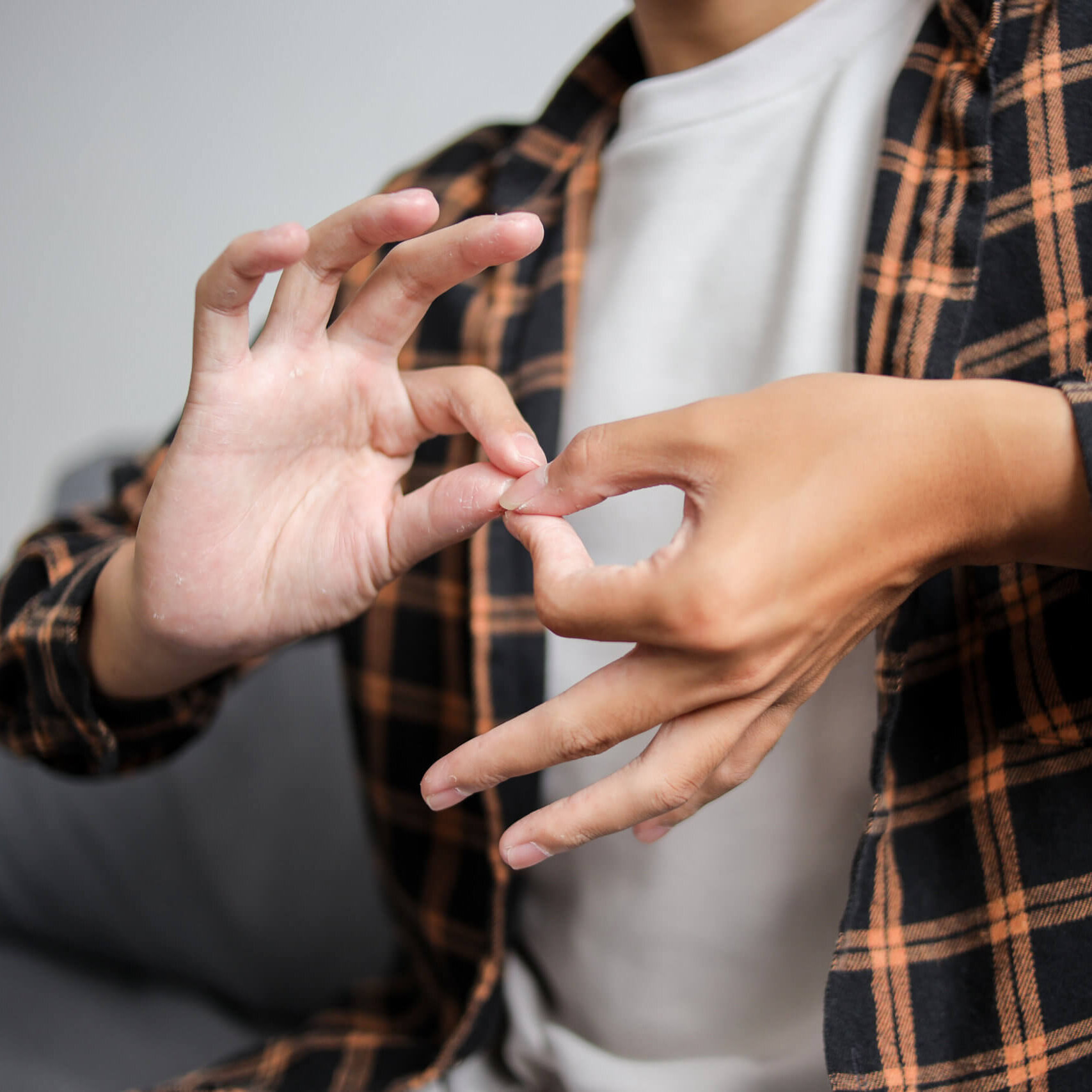 A person wearing a black and orange plaid flannel shirt over a white t-shirt uses both hands to form the ASL sign for 'interpreter,' photographed from the chest down against a blurred gray sofa background.